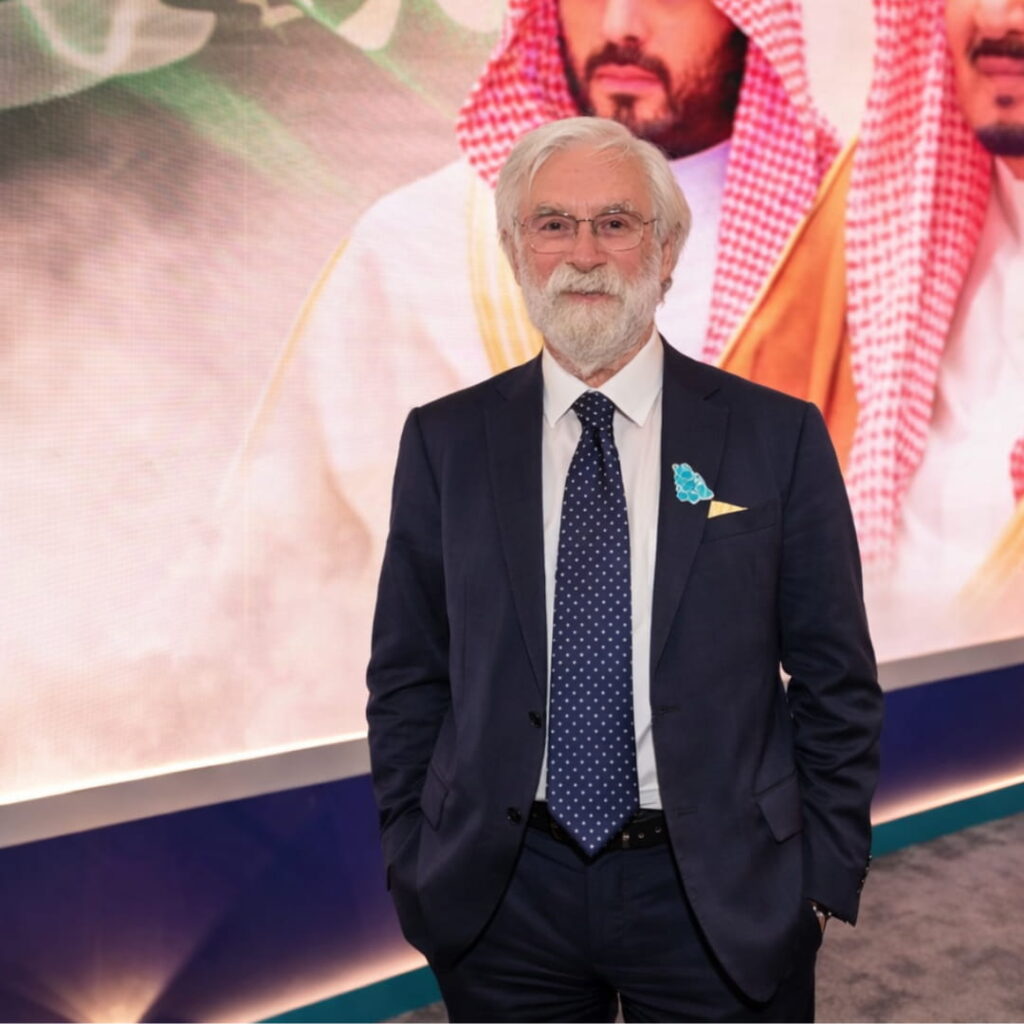 Roy standing at an event in a navy suit and tie with a conference display in the background.