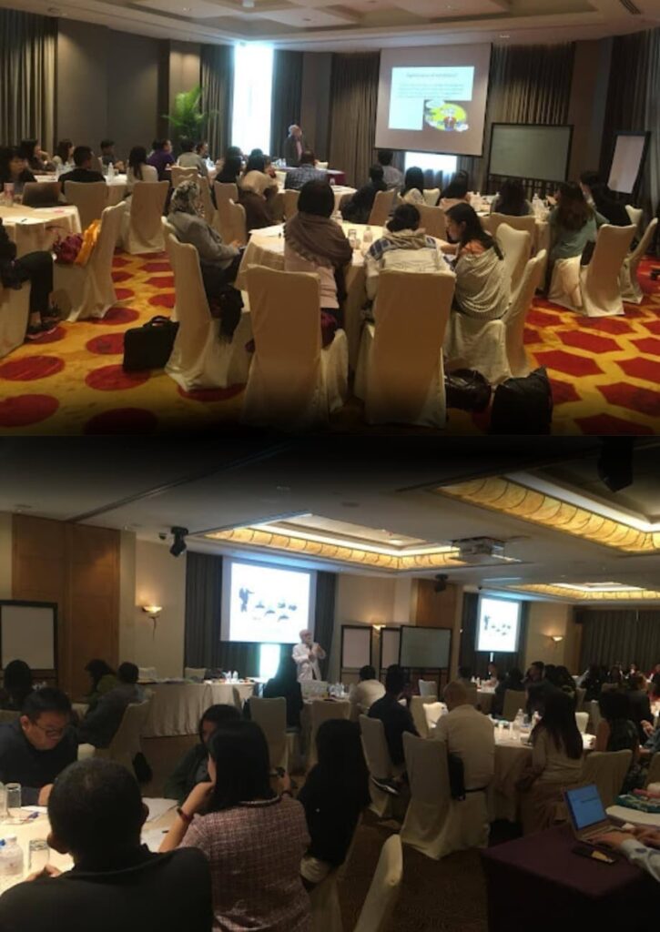 Conference attendees seated at round tables while a speaker presents on a screen at the front of the room.