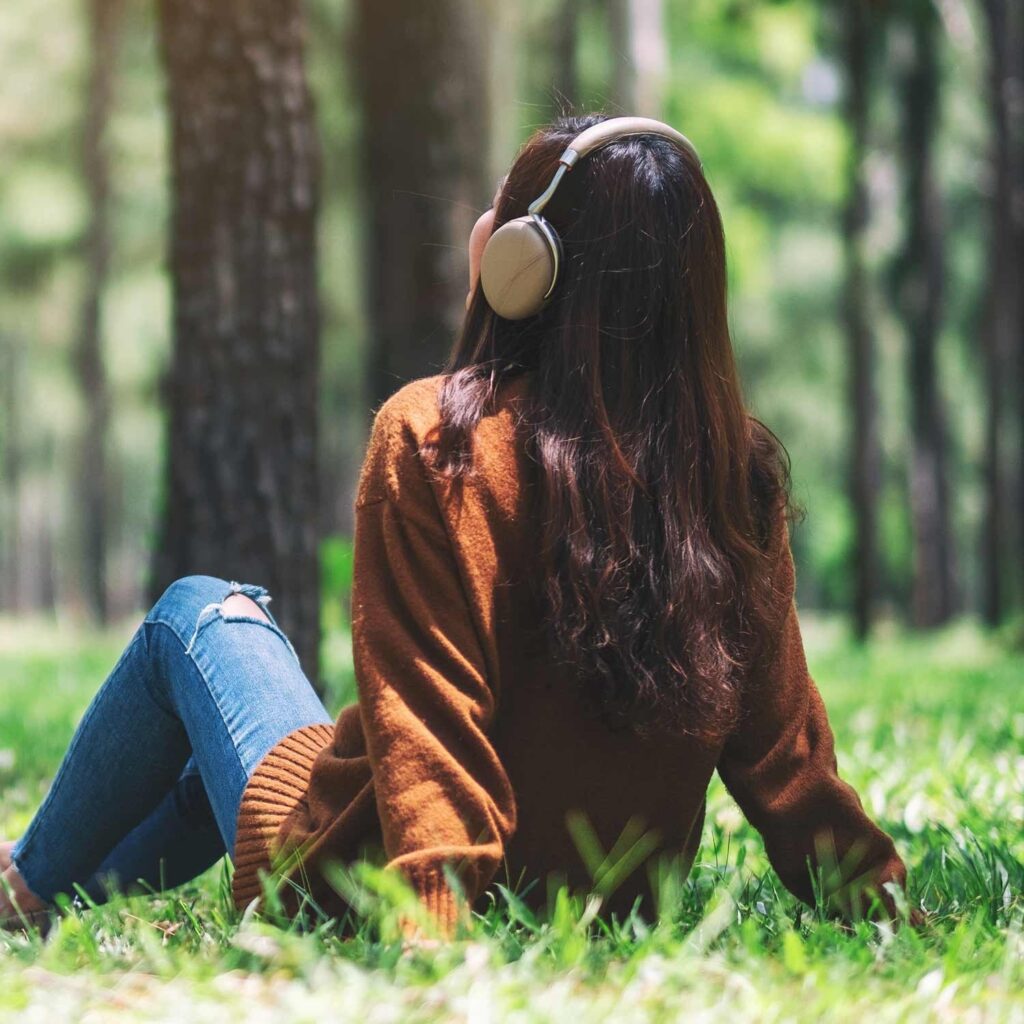 Woman sitting on grass in a forest wearing headphones, relaxing and listening to audio content