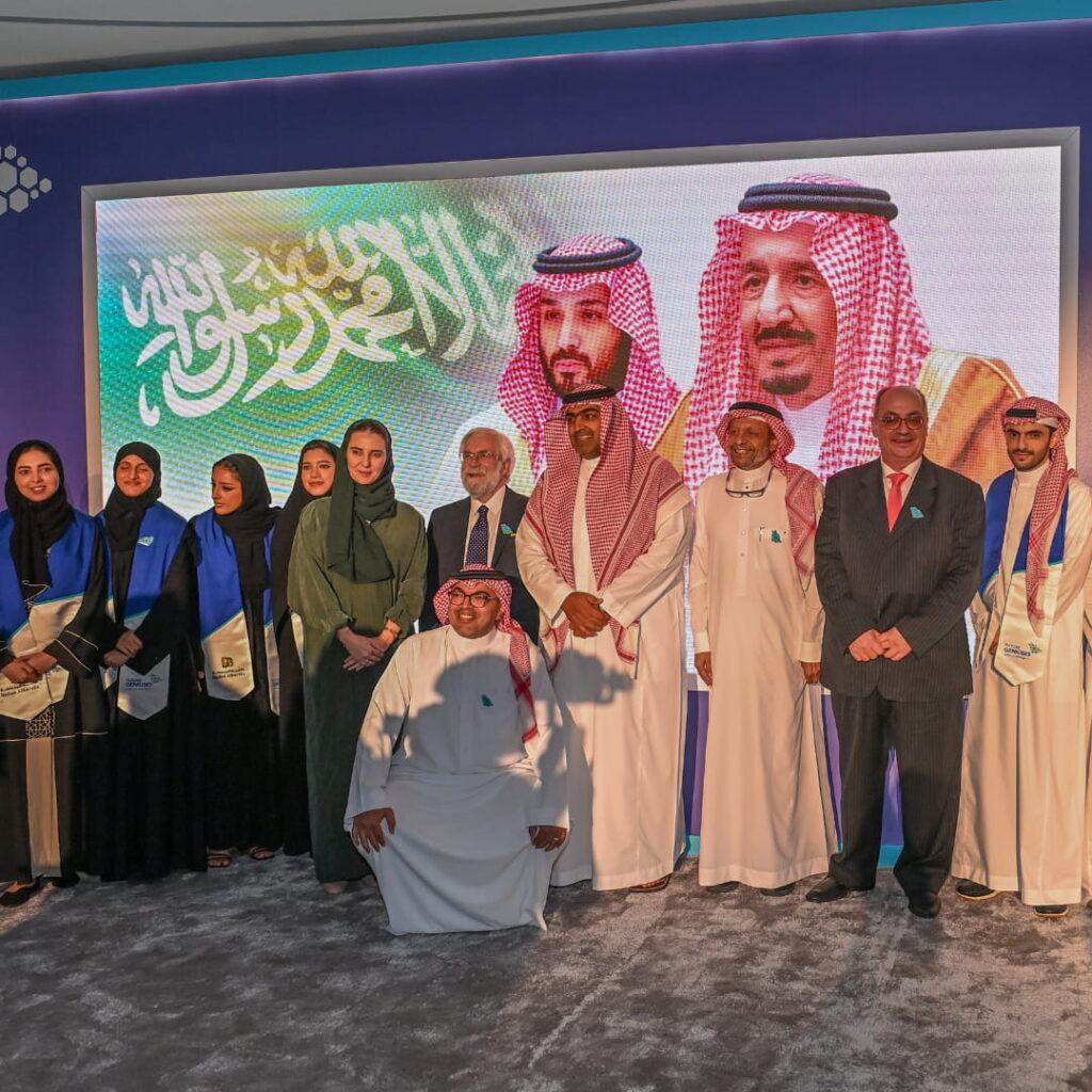 Group of professionals posing on stage at a formal event with a large Saudi-themed display in the background.