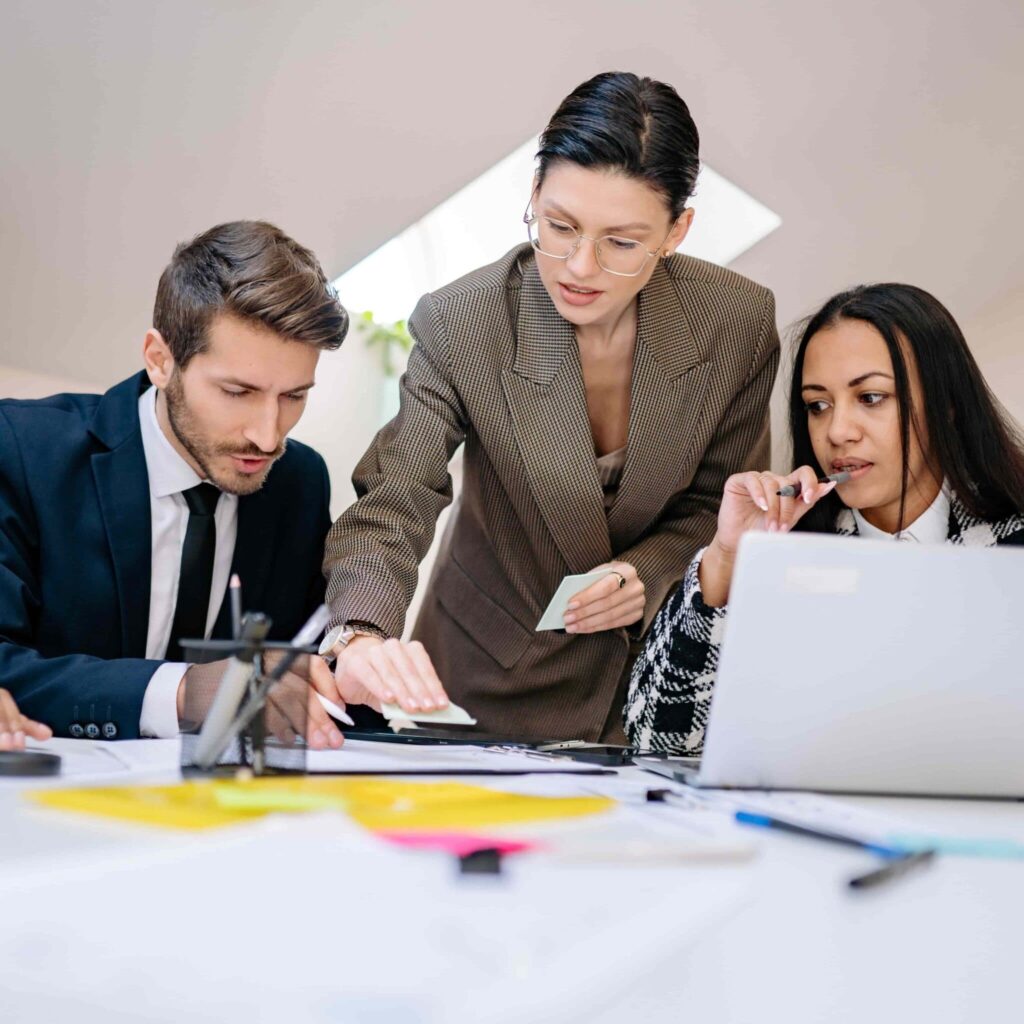 Three professionals collaborating at a desk while reviewing documents and working on a laptop.