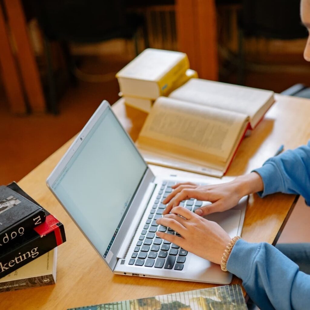 Person using a laptop while reading books and studying online resources.